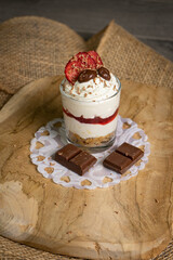 A delicious dessert in a glass with a crunchy biscuit base, smooth cream cheese layer, and strawberry jam. Topped with grated chocolate, dried strawberries, and chocolate candies, served on a rustic w