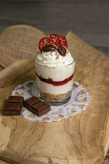 A delicious dessert in a glass with a crunchy biscuit base, smooth cream cheese layer, and strawberry jam. Topped with grated chocolate, dried strawberries, and chocolate candies, served on a rustic w
