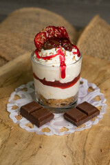 A delicious dessert in a glass with a crunchy biscuit base, smooth cream cheese layer, and strawberry jam. Topped with grated chocolate, dried strawberries, and chocolate candies, served on a rustic w