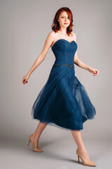 Naklejka premium Beautiful redhead woman walking in studio, sensual portrait. Deep blue sweetheart neckline midi dress. Corset sleeveless classic evening gown, festive outfit. Touching look for an event, flying skirt