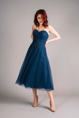 Fototapeta premium Cheerful redhead woman dancing in studio, smiling portrait. Deep blue sweetheart neckline midi dress. Corset sleeveless classic evening gown, festive outfit. Touching look for an event, flying skirt