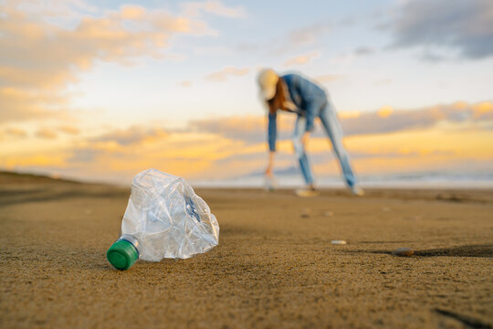 Global environmental pollution. A female volunteer collects plastic bottles on the ocean shore. Cleaning of the coastal zone. Sunset in the background