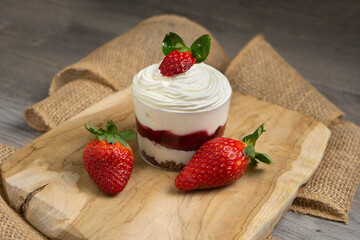 a dessert placed on a wooden board. It appears to be a layered parfait or cheesecake in a small glass, topped with whipped cream and a strawberry. Two whole strawberries are next to the dessert on the