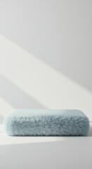 A plush blue fur cushion rests on a bright surface, illuminated by natural light creating soft shadows. The minimalistic setting enhances the cushion's textures