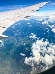 Obraz premium An airplane wing dominates the frame against a backdrop of fluffy white clouds and a patchwork of green and brown fields, seen from above through the plane's window.