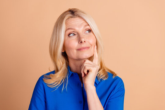 Portrait of a thoughtful mature woman in a blue shirt on beige background, touching her face and expressing curiosity and contemplation