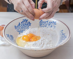 Cracking an egg with your hands and pouring it  over flour in a ceramic bowl.