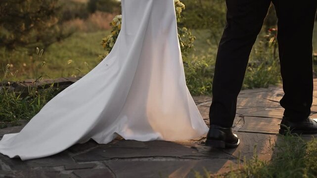 Bride in long dress with hem and groom in black shoes and trousers walk along path among flowers and grass in the summer at sunset