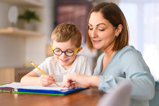 homework and school concept - smiling student boy with book writing to notebook at home with his mother help her