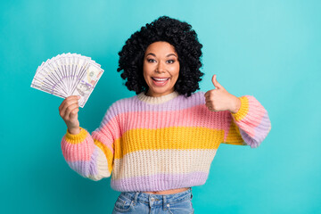 Joyful young woman holding money while posing with thumbs up, showcasing happiness and financial success