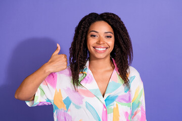 Stylish young woman wearing a colorful shirt gives a thumbs up, smiling against a vibrant purple...