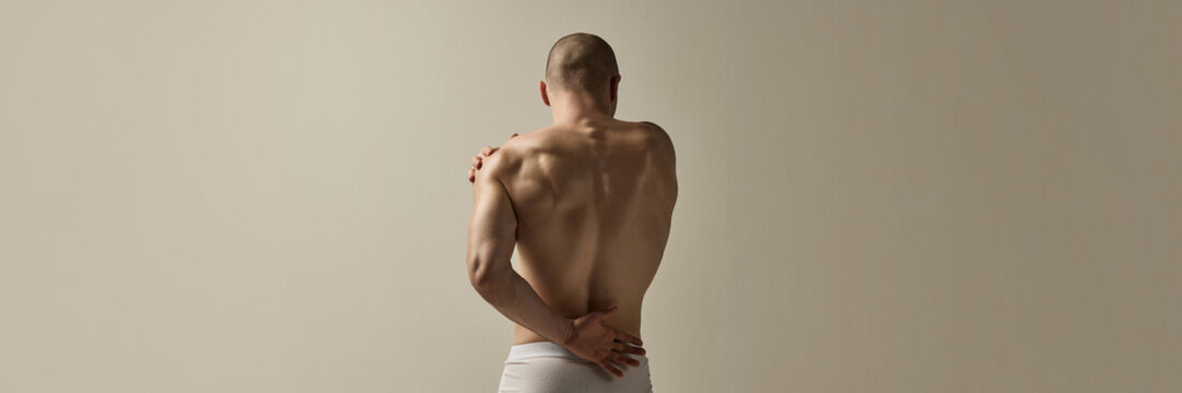 Banner. Masculine man, dressed underwear, standing with back arched and arms crossed in emotional movement against soft- beige studio background. Concept of beauty, fashion, style, masculinity.