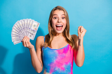 Joyful young woman happily holds dollar bills in a colorful outfit against a vibrant blue...