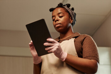 Chest up shot of adult Black female chef in apron using digital tablet while cooking following recipe in bakery kitchen, copy space