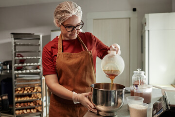 Medium shot of young female baker wearing hairnet pouring maple syrup into metal bowl mixing...