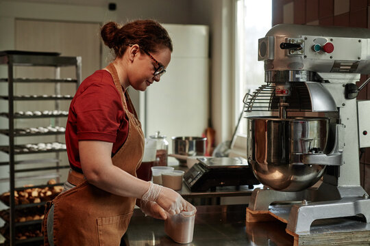 Side view of young female baker wearing apron adding tablespoon of ingredient into stainless steel bowl of large dough kneading machine, while cooking in commercial kitchen