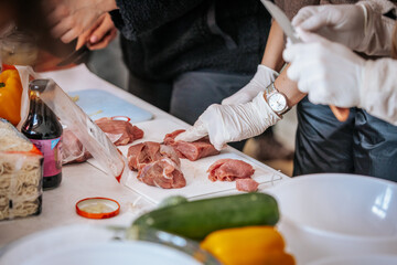 Close-up of hands wearing gloves cutting raw meat on a white cutting board. The scene suggests food preparation, hygiene, and cooking, with a watch visible on one wrist.
