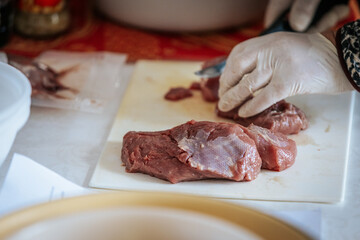 Close-up of hands wearing gloves cutting raw meat on a white cutting board. The scene suggests food preparation, hygiene, and cooking, with a watch visible on one wrist.
