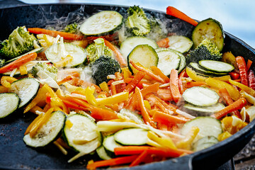 A steaming vegetable stir-fry with zucchini, bell peppers, carrots, and broccoli is being cooked in a pan. A spatula is stirring the fresh, colorful ingredients.