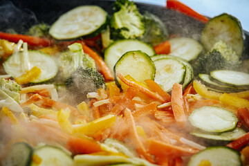 A steaming vegetable stir-fry with zucchini, bell peppers, carrots, and broccoli is being cooked in a pan. A spatula is stirring the fresh, colorful ingredients.