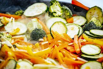 A steaming vegetable stir-fry with zucchini, bell peppers, carrots, and broccoli is being cooked in a pan. A spatula is stirring the fresh, colorful ingredients.
