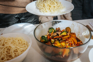 A group of people serves stir-fried beef with vegetables and noodles onto plates during a shared meal. The dish is rich in colors and flavors, reflecting Asian cuisine.