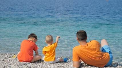 Father and two children sit on beach relaxing, playing and throwing pebbles