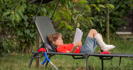 Child with leg in cast lying on garden lounger, relaxing by reading a book