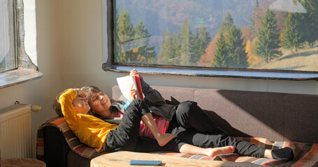 Mother and son lying on sofa reading a book in a mountain rustic chalet