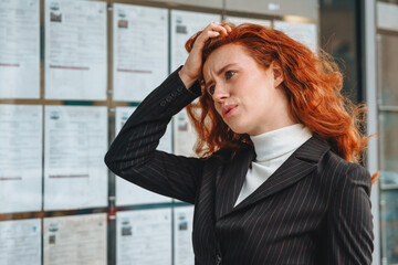 Fototapeta premium Woman with red hair appears stressed while reviewing listings on information board at advertised properties, trying to find house to buy or rent on bulletin board Outside Building
