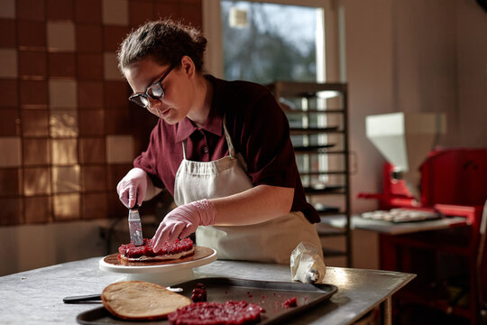 Medium shot of young female confectionary chef enjoying process of assembling delicious cake spreading cherry jam over moist layer with knife, while working at table in patisserie kitchen