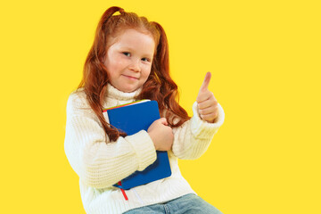 Cheerful girl with red hair gives thumbs up while holding a book against a yellow background
