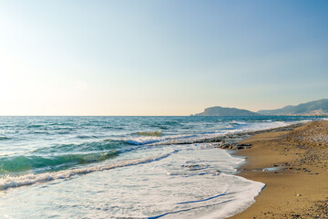 View of a serene sandy beach with gentle waves under the warm light of the setting sun during summer.