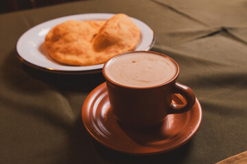 Coffee in a brown cup next to cooked dough on a tablecloth.