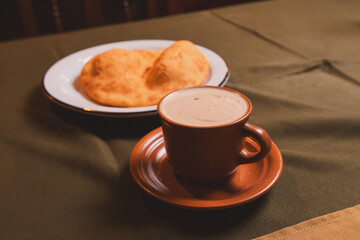 Coffee in a brown cup next to cooked dough on a tablecloth table.