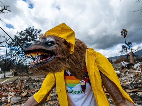 Wolf mascot wearing a yellow raincoat stands beside an "Altadena Not For Sale!" sign amid Eaton Fire destruction in Altadena, California, highlighting community resilience after wildfire