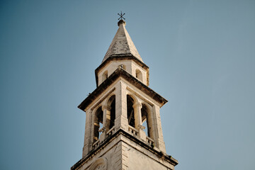 Historic church bell tower with a pointed spire in old town Budva set against a clear blue sky. The aged stone architecture and intricate details reflect timeless elegance and religious heritage.