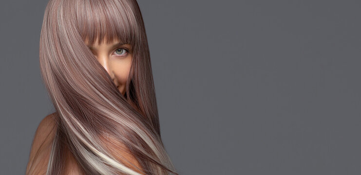 Woman with long, silky, ash brown flying hair with platinum highlights covering part of her face, looking at the camera with a soft expression against a gray background..