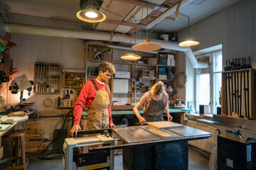 Handyman overseeing novice woman joiner working on circular saw in woodworking workshop, ready to assist and guide. Collaboration for learning in carpentry, manual labor, board repair and cutting