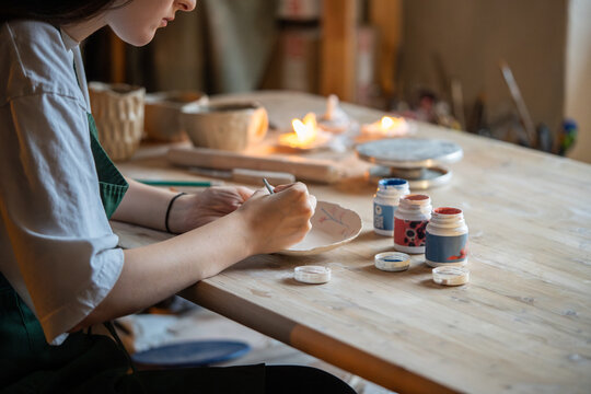Decorating pottery. Woman ceramist holding paintbrush and drawing cute ornament on clay plate. Creative classes of work with earthenware, creating an authors style in tableware design. Artist at work.
