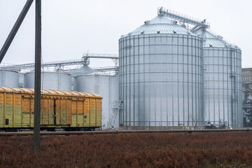 Grain silos tower above a railway line as yellow freight cars stand empty against a cloudy backdrop...