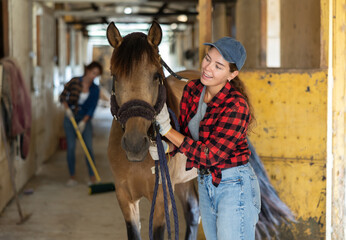 Female horse farm worker takes horse out of stall, holds animal by bridle and leads pet for walk. Woman and horse before walk and training trip.