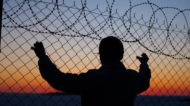Refugee at the border. Silhouette of a refugee woman in a baseball cap at sunset, clinging to a fence with barbed wire. Concept of hope, freedom and a better life