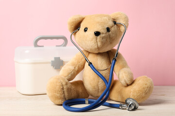 Pediatrics concept. Teddy bear with stethoscope and first aid kit on wooden table against pink...