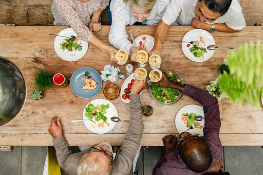 Diverse senior friends enjoying meal together, toasting drinks. Diverse elderly toasting drinks, eating healthy meal at wooden table. Healthy dining, diverse gathering. Healthy seniors lifestyle - Powered by Adobe
