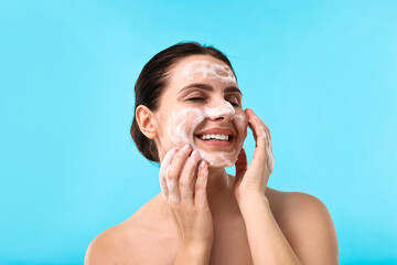 Smiling woman washing her face with cleansing foam on light blue background