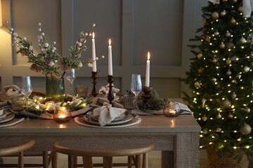Christmas table setting with stylish dishware, glasses and burning candles in festive decorated room