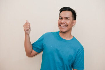 A Southeast Asian man in a turquoise green tshirt smiling and pointing thumbs towards up right in a...