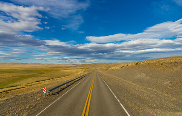 A long, empty road stretches endlessly through a vast, arid landscape under a bright blue sky with scattered clouds. The scene evokes adventure, freedom, and solitude, perfect for travel exploration