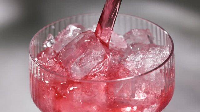 fresh red drink pouring into a cocktail glass with ice cubes on a gray sunny restaurant tabletop, slow motion